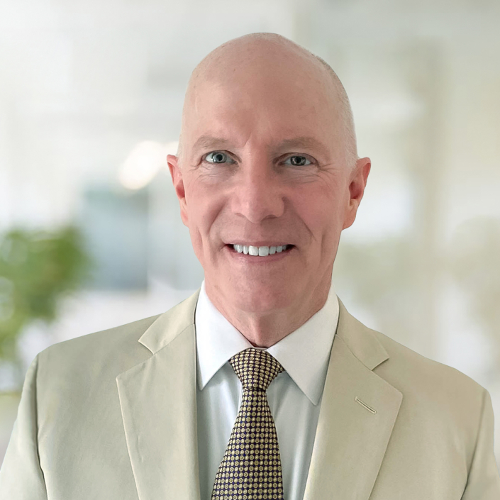 Tom Holleran wearing a cream-colored sport coat and tan printed tie. An office is visible in soft focus behind him.