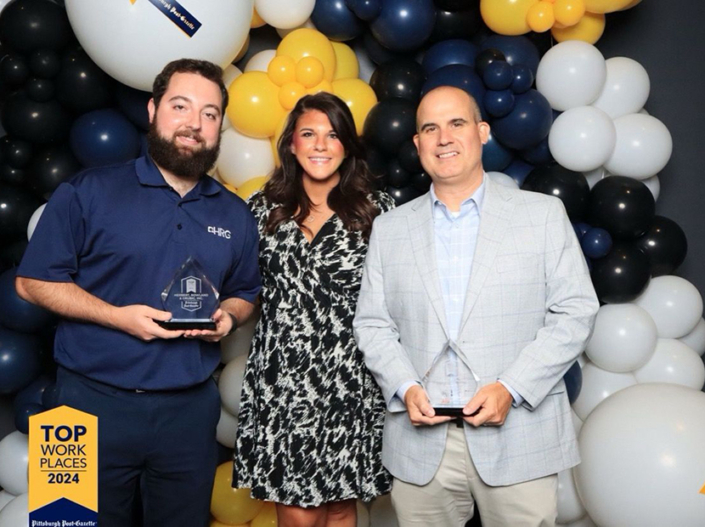 Clayton Whitlatch, Kara Smith, and Andrew Scampone accept HRG's awards at the Pittsburgh Top Workplaces ceremony, September 2024. They are standing in front of a wall of navy, yellow, and white balloons.