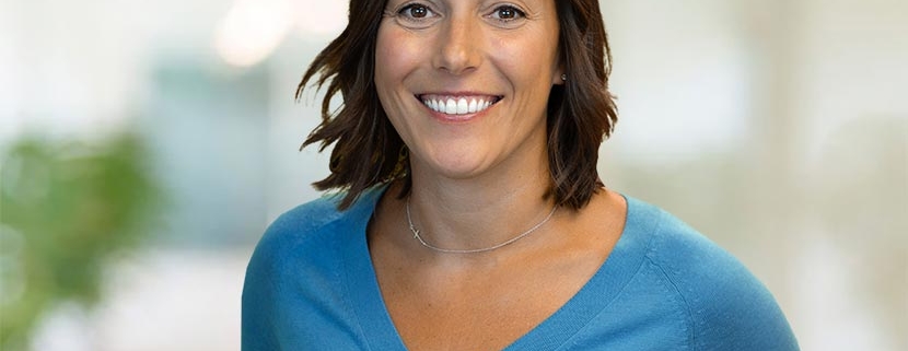 Photo of Erin Threet wearing a light blue, v-neck sweater and standing in front of a blurred office background